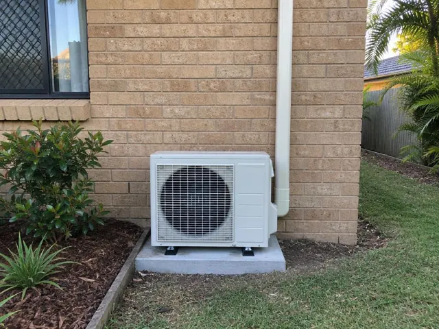 Outdoor air conditioner unit on a concrete pad beside a brick house wall in Carrara
